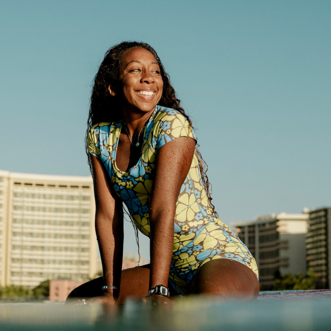 Woman in a floarl surfsuit sitting on a surfboard in the ocean with buildings in the background.