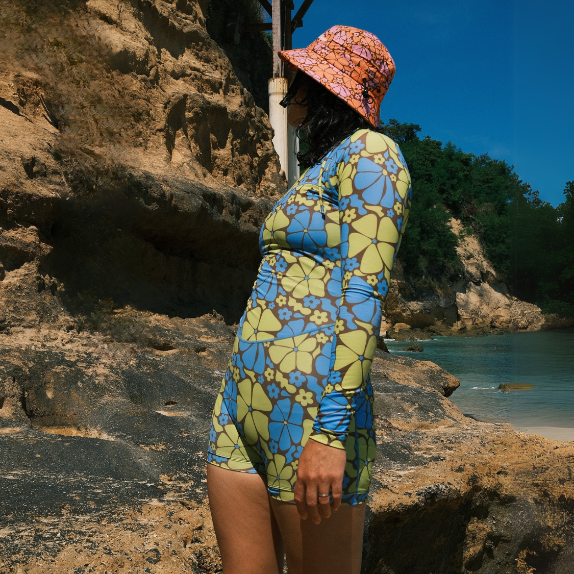 Person in floral swimsuit and hat standing on a rocky beach.