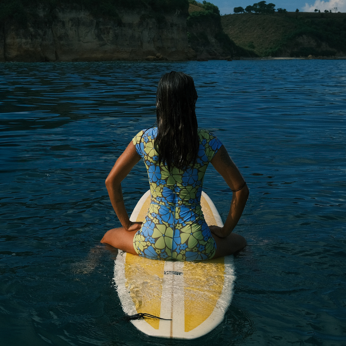 Woman in floral sun-smart swimsuit sitting on a surfboard in the ocean.
