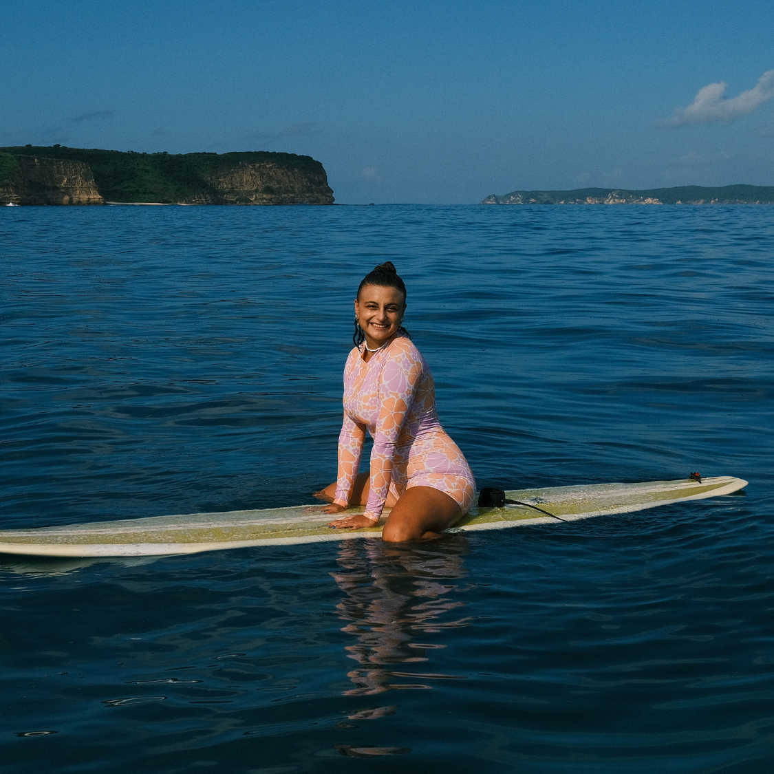 Woman in a pink swimsuit sitting on a surfboard in the ocean with cliffs in the background